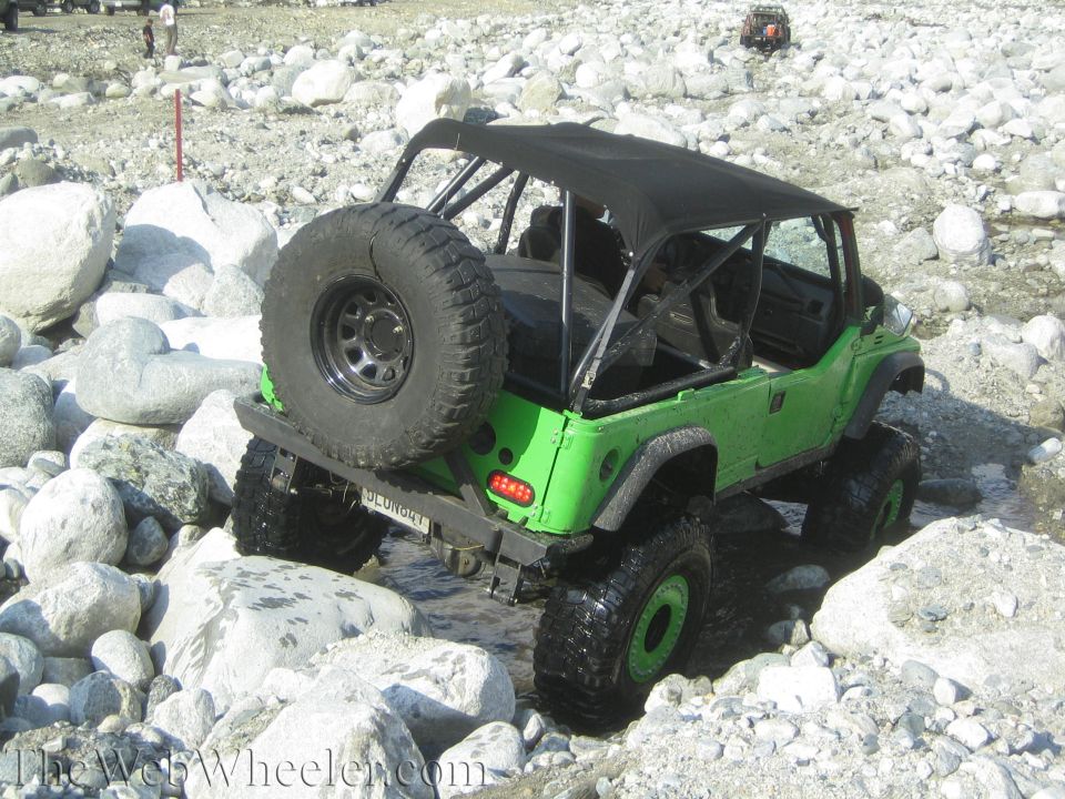 Green 1987 Suzuki Samurai with a spare tire on the back, driving over rocks in a stream