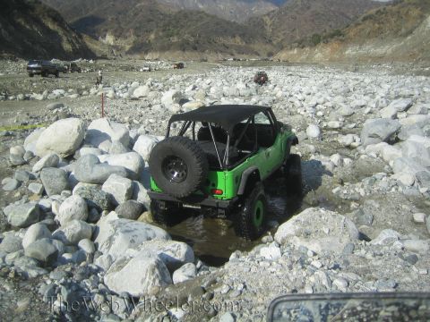 1987 Suzuki Samurai - A bright green Jeep Wrangler drives through a rocky riverbed, with other vehicles visible in the distance.