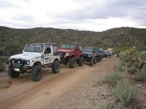 1987 Suzuki Samurai - A convoy of off-road vehicles travels down a dusty desert trail under a cloudy sky.