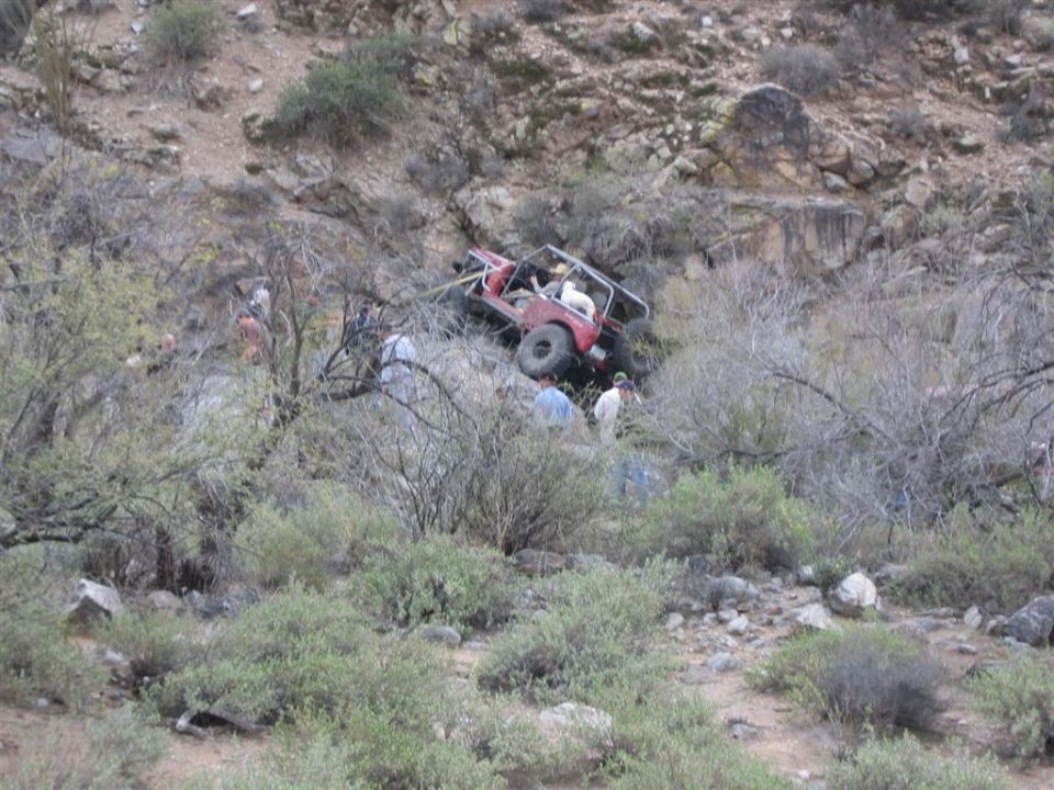 Red 1987 Suzuki Samurai off-road vehicle tilted on a rocky hillside with people around it