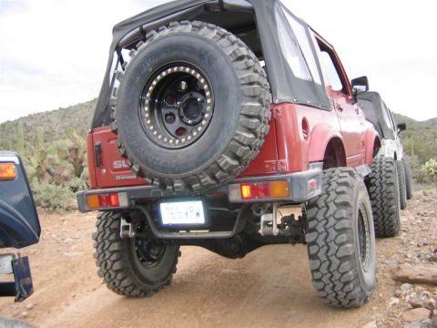 1987 Suzuki Samurai - A red 1987 Suzuki Samurai is seen from the rear, parked with other vehicles in a desert landscape.