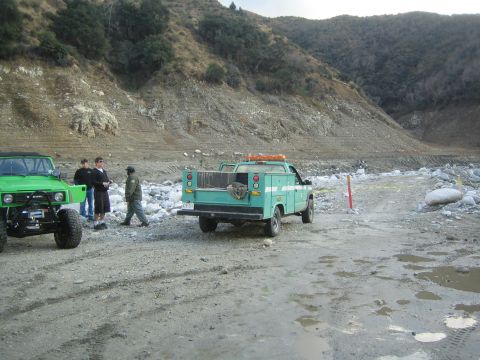 1987 Suzuki Samurai - Two vehicles are parked on a dirt path in a rocky, dry riverbed with a steep, barren hillside in the background.
