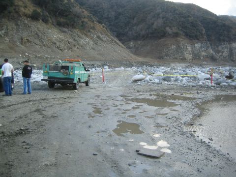 1987 Suzuki Samurai - A teal utility truck is parked on a muddy, rocky area near a hillside, with two people standing nearby.