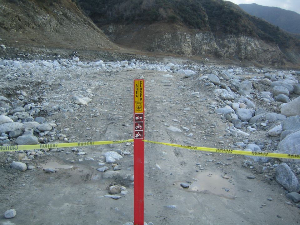 Sign says 'AREA CLOSED' with icons for motorcycles and ATVs. Yellow caution tape blocks a rocky, muddy path