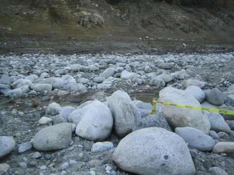 1987 Suzuki Samurai - Caution tape is visible across a rocky riverbed, with a dirt embankment in the background.