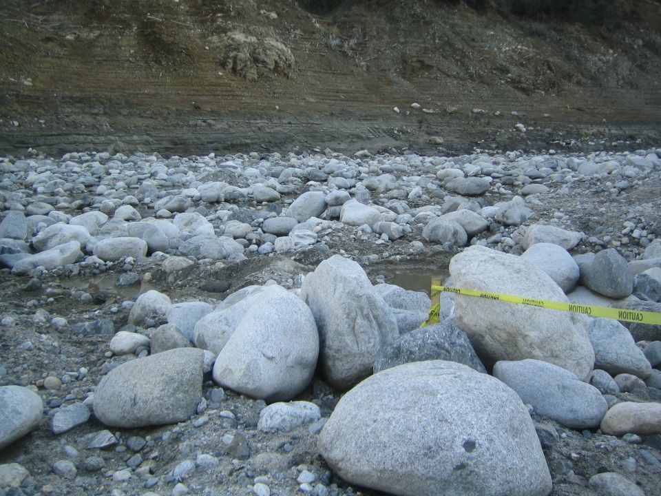 caution tape across a rocky riverbed with a dirt embankment in the background