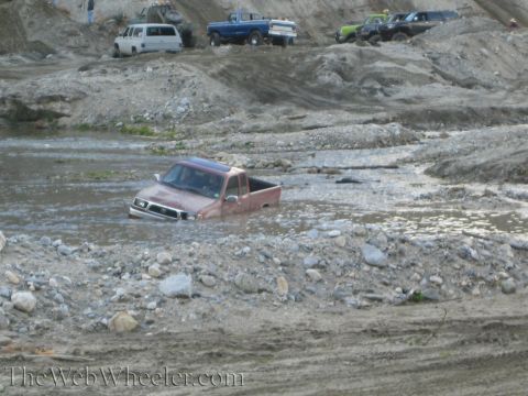 1987 Suzuki Samurai - Multiple off-road vehicles navigate a muddy, watery terrain, with one red pickup truck partially submerged.