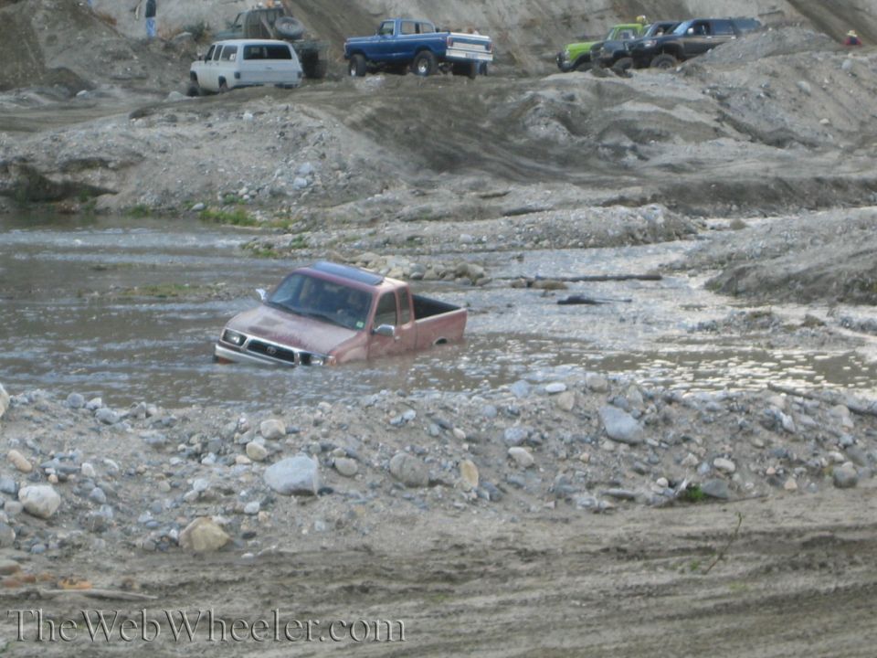 Red pickup truck stuck in water, other off-road vehicles visible