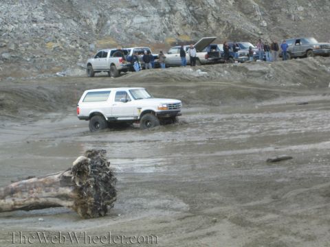 1987 Suzuki Samurai - A white Ford Bronco navigates a muddy terrain while other vehicles are parked on the embankment above.
