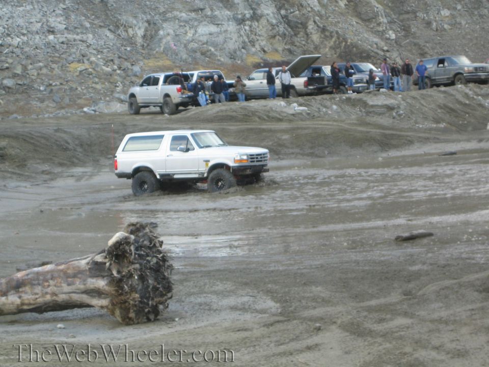 White Ford Bronco driving through mud with other trucks parked on a hill