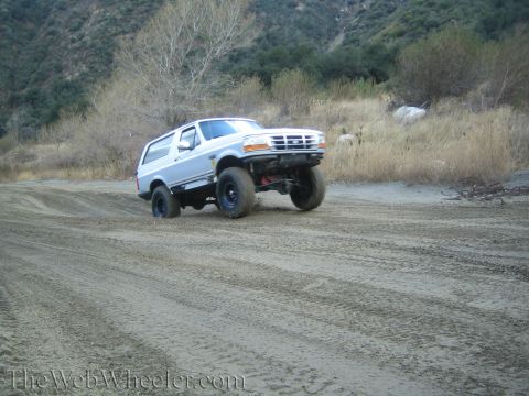 1987 Suzuki Samurai - A silver Ford Bronco navigates a sandy terrain with dry brush and hills in the background.