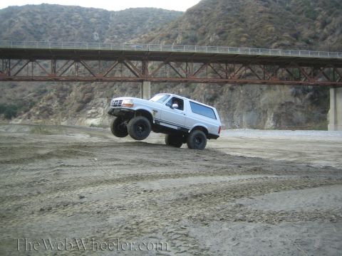 1987 Suzuki Samurai - Silver SUV with lifted suspension airborne over dirt, with a bridge in the background.