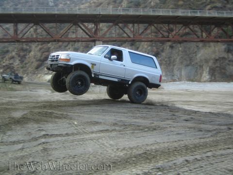 1987 Suzuki Samurai - Ford Bronco airborne over a dirt track, with a bridge overhead and another vehicle in the distance.