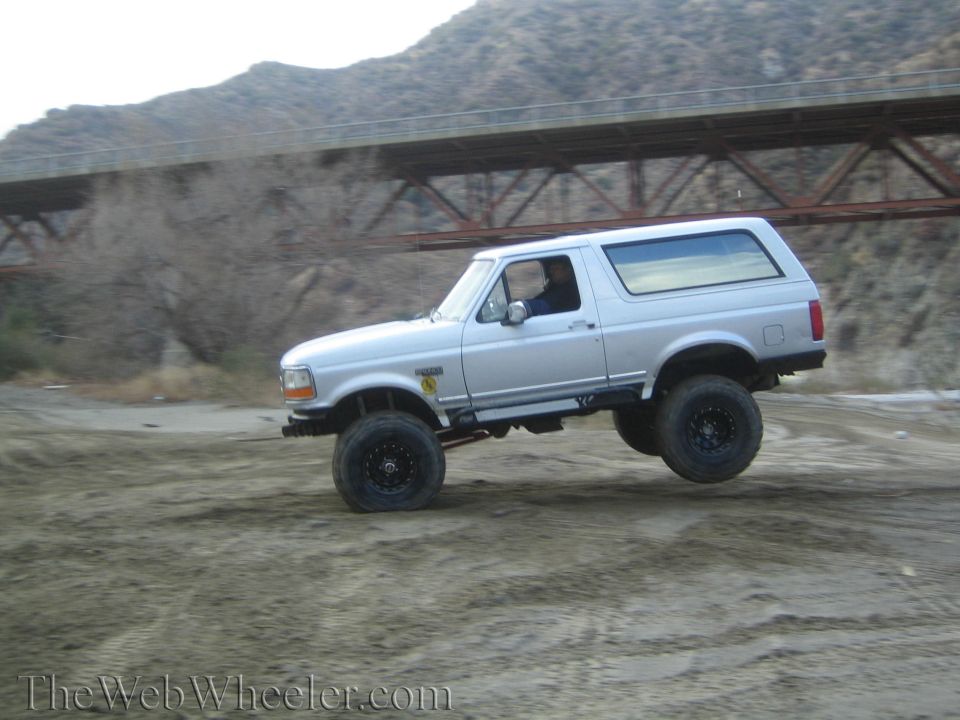 Silver Ford Bronco with lifted suspension driving off-road. It has large tires and a roll bar visible inside