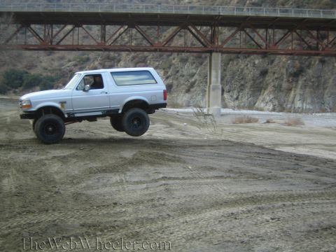 1987 Suzuki Samurai - Silver Ford Bronco with lifted suspension driving off-road under a bridge.