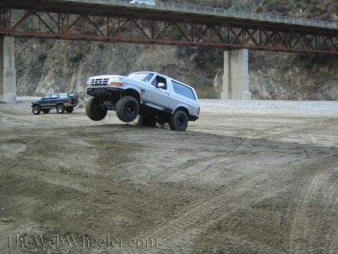 1987 Suzuki Samurai - A lifted Ford Bronco jumps over a dirt mound near a bridge, with another SUV parked nearby.