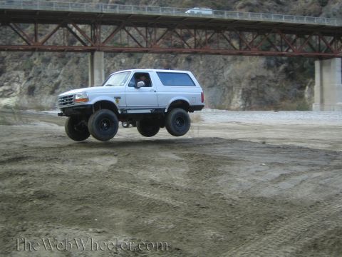 1987 Suzuki Samurai - White Ford Bronco airborne over dirt with a bridge overhead.