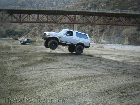 1987 Suzuki Samurai - A silver SUV is captured mid-air over a dirt terrain, with a large bridge structure in the background.