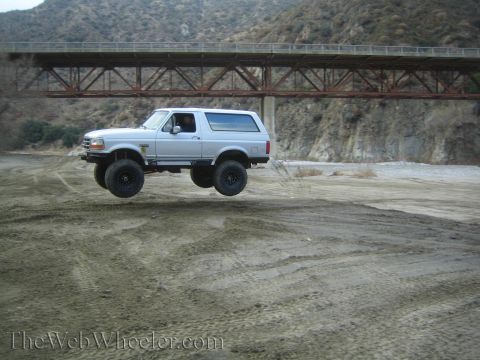 1987 Suzuki Samurai - A silver Ford Bronco airborne over a dry riverbed, with a large bridge overhead.