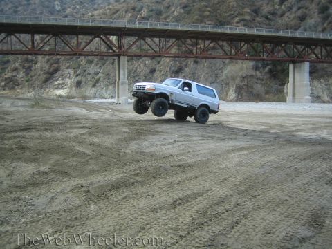 1987 Suzuki Samurai - Silver Ford Bronco airborne over dirt, front wheels off the ground, under a bridge.