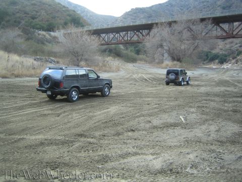 1987 Suzuki Samurai - Two SUVs, a dark SUV and a Jeep, are parked in a dry riverbed with a bridge overhead.