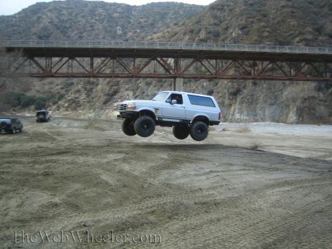 1987 Suzuki Samurai - Ford Bronco airborne over a dry riverbed with a bridge overhead.