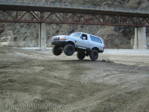 1987 Suzuki Samurai - A lifted white Ford Bronco performs a jump in a dry riverbed beneath a large bridge.