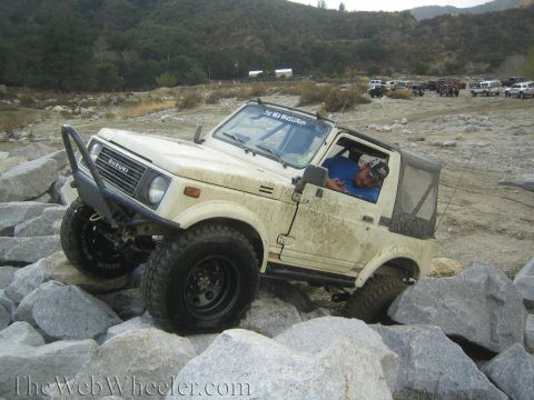 1987 Suzuki Samurai - A man drives a cream-colored 1987 Suzuki Samurai over large rocks in a dry riverbed with other vehicles in the background.