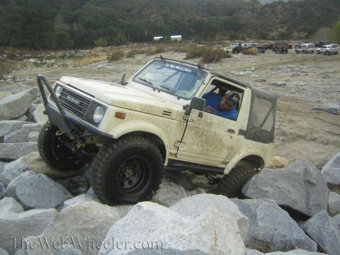 1987 Suzuki Samurai - A white 1987 Suzuki Samurai is shown off-roading over large rocks, with other vehicles visible in the background.