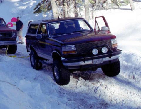 1993 Ford Bronco - A dark blue Ford Bronco is stuck in the snow, with a red truck nearby.