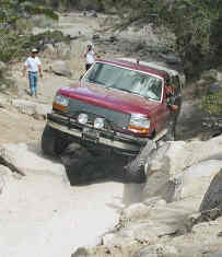 1993 Ford Bronco - A red Ford Bronco tackles a challenging off-road course with two people visible inside.