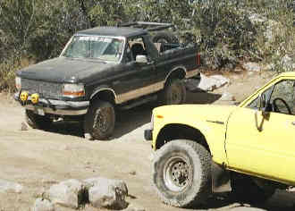 1993 Ford Bronco - Two SUVs, a black Ford Bronco and a yellow pickup, navigate a dusty trail.