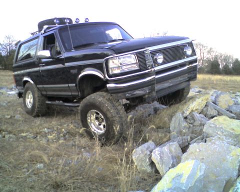 1995 Ford Bronco - This black 1995 Ford Bronco is equipped with a roof rack and off-road tires, navigating a rocky terrain.
