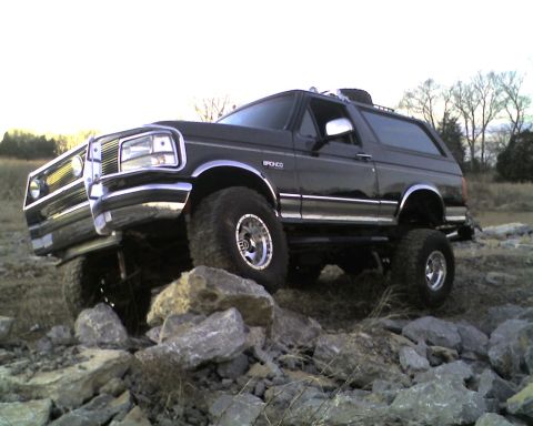 1995 Ford Bronco - Lifted black Ford Bronco crawling over a pile of broken limestone rocks, shot from a low front three-quarter angle so the front tire is lif…