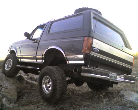 1995 Ford Bronco - Low three-quarter rear view of a black lifted Ford Bronco articulating over rocks, with the rear axle flexed and the polished wheels showin…