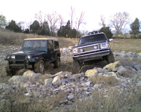 1995 Ford Bronco - Two 4x4s parked side by side on a rocky, brush-covered field in winter: a dark blue Jeep Wrangler YJ with a tan soft top, square headlights…