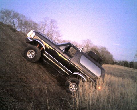 1995 Ford Bronco - Lifted black Ford Bronco climbing a steep dirt embankment, viewed from the passenger side against a clear blue sky and bare winter trees. C…