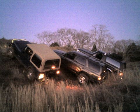1995 Ford Bronco - Dusk shot, tilted at a steep angle, of three 4x4s lined up on a grassy hillside with their lights on: a Jeep Wrangler with a tan hardtop, a…