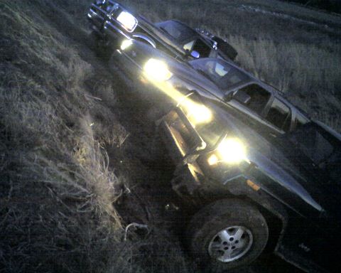1995 Ford Bronco - Grainy night shot, tilted at a steep angle, of a dark Ford Bronco with headlights and roof-mounted driving lights blazing while parked in a…
