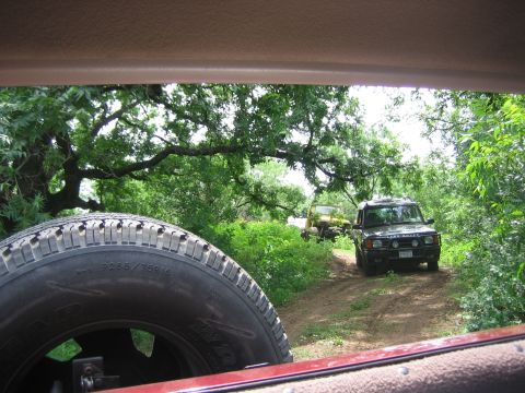 1996 Ford Bronco - Two SUVs navigate a dirt path through a wooded area, with a spare tire visible in the foreground.