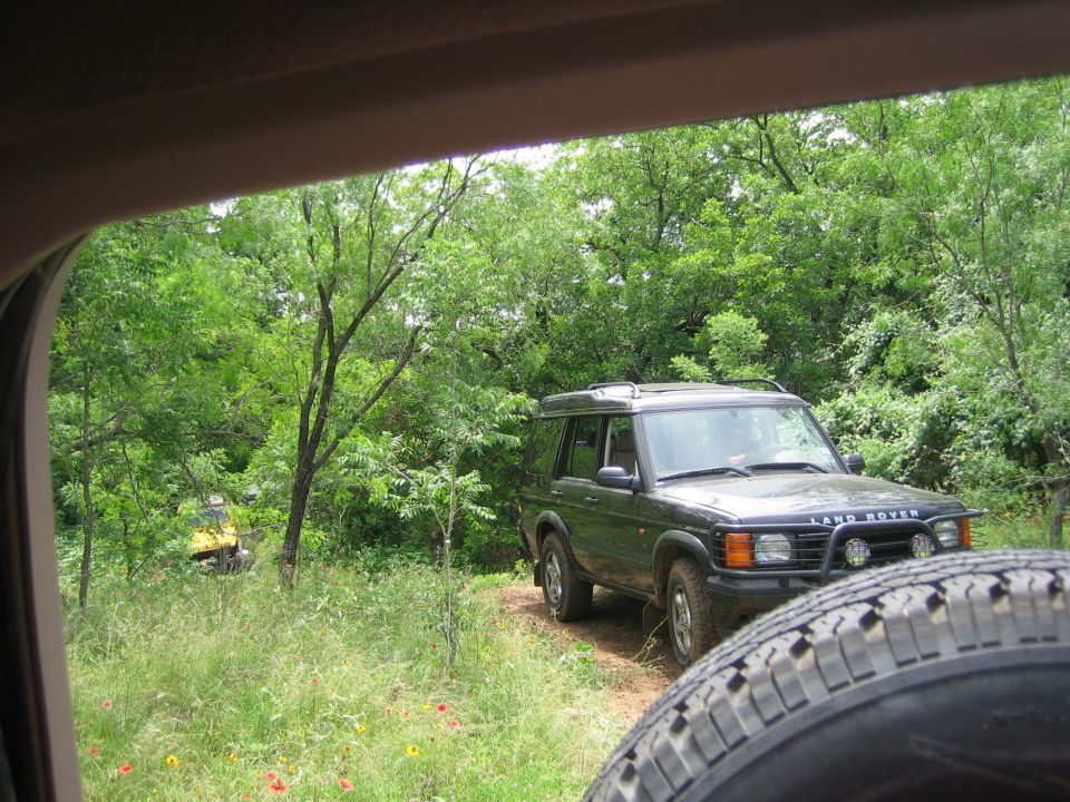 Dark green Land Rover Discovery parked on a dirt path, with a yellow vehicle visible in the background trees. Spare tire in foreground
