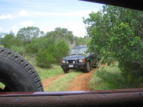 1996 Ford Bronco - Land Rover Discovery navigates a dirt path surrounded by greenery under a partly cloudy sky.