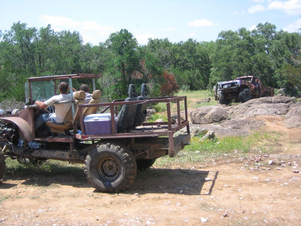 Side view of a custom off-road vehicle with a roll cage and open rear bed, another off-road vehicle climbs rocks in background