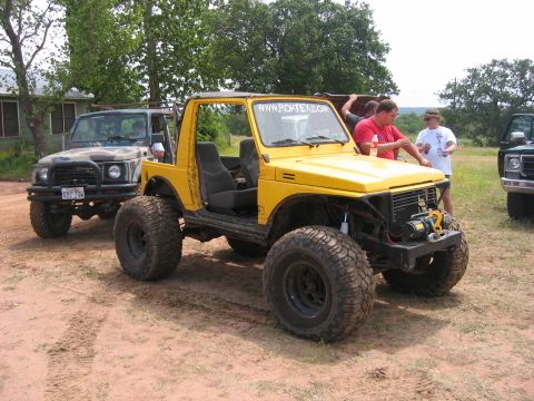 1996 Ford Bronco - Several off-road vehicles are parked on a dirt lot with trees and a building in the background.
