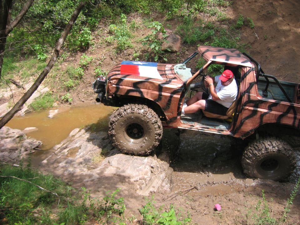 1996 Ford Bronco with Texas flag on hood, driving off-road through mud and water