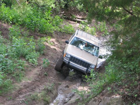1996 Ford Bronco - A Jeep Cherokee XJ navigates a muddy, off-road trail with dense foliage on either side.