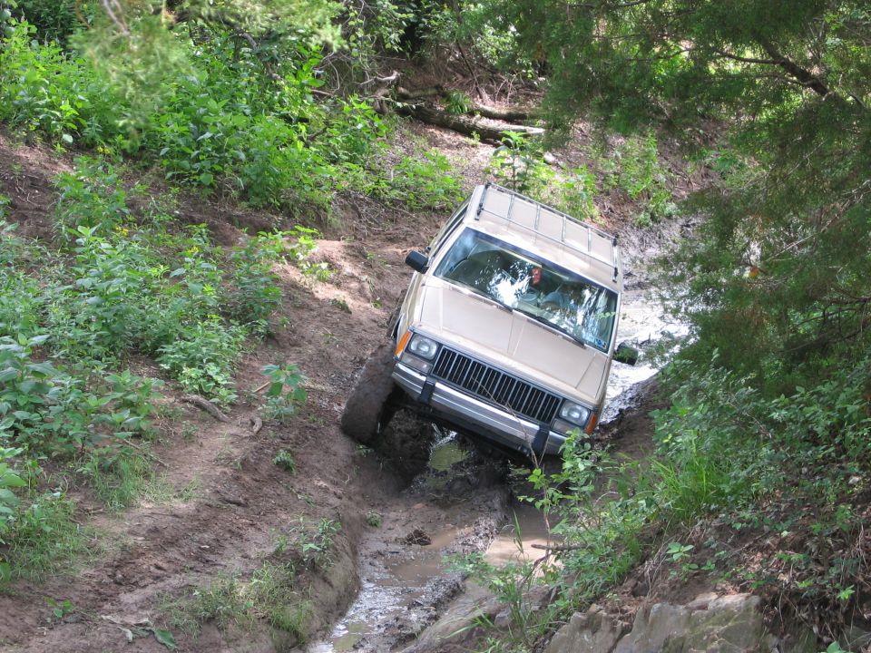 tan Jeep Cherokee XJ driving through mud and water on a wooded trail