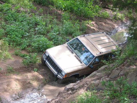 1996 Ford Bronco - A tan Jeep Cherokee XJ navigates a challenging off-road course, getting stuck in a muddy water-filled ditch.