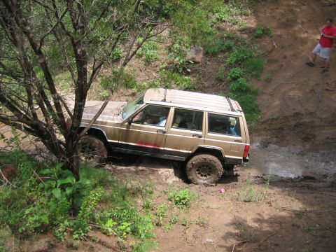 1996 Ford Bronco - A tan Jeep Cherokee XJ tackles a challenging muddy off-road course, with one person observing from the bank.