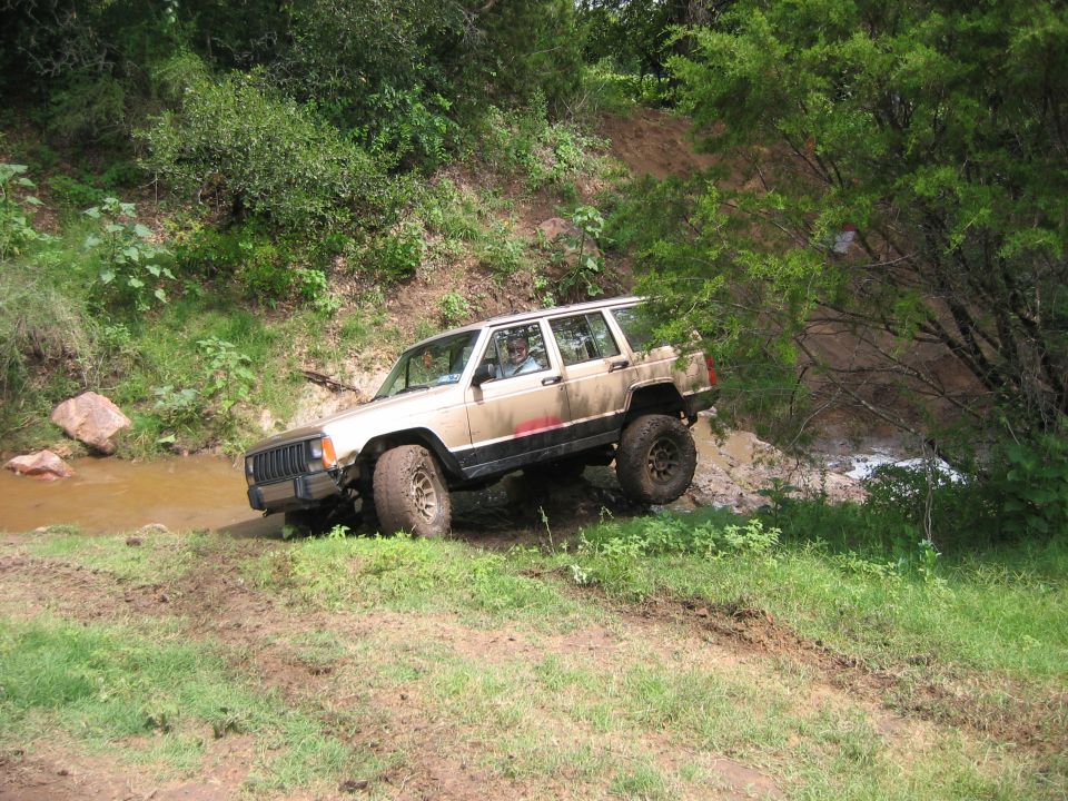Tan Jeep Cherokee XJ with lifted suspension driving through muddy terrain near a creek